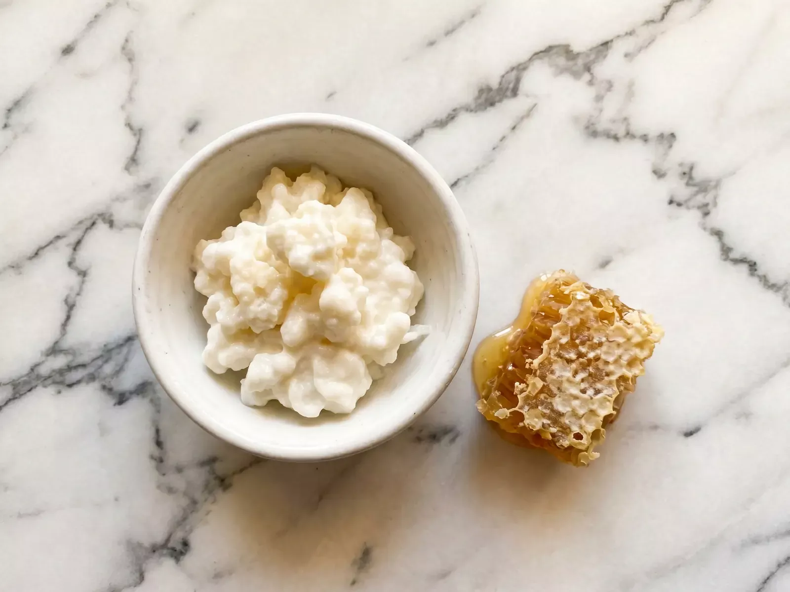 Ciotola di kefir naturale con panal de miel accanto, su marmo venato con luce calda dall&rsquo;alto.