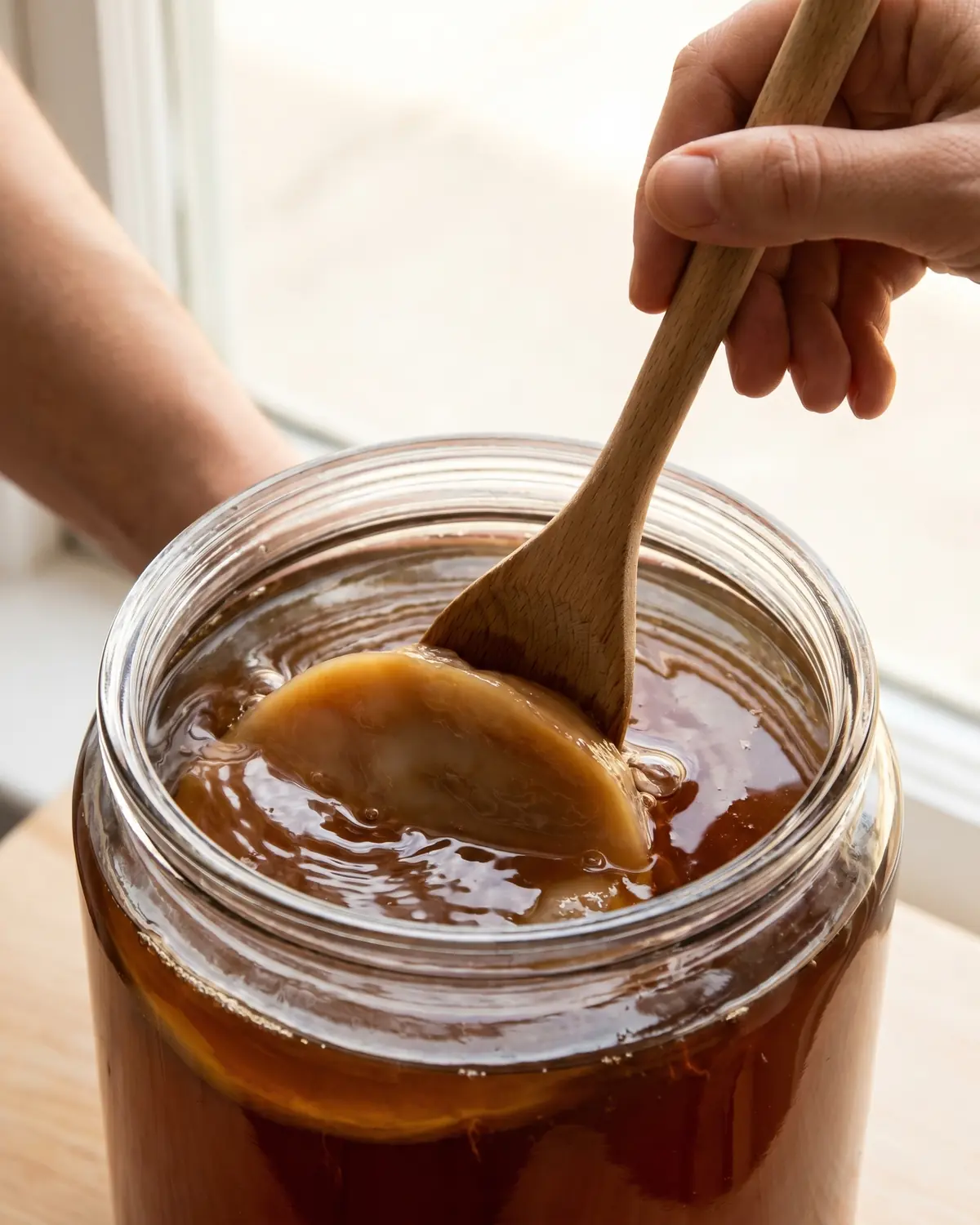 kombucha lidl Cucchiaio di legno che spinge delicatamente lo SCOBY sotto il tè ambrato durante la preparazione.
