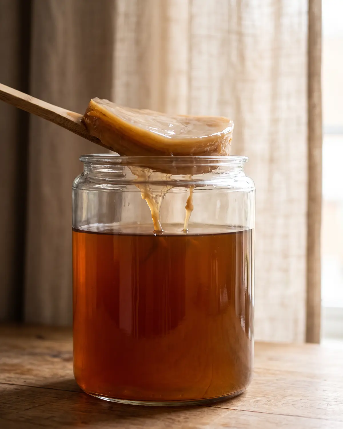scoby hotel Primo piano di un vaso con SCOBY e filamenti morbidi, accanto a una tenda di lino sfocata.