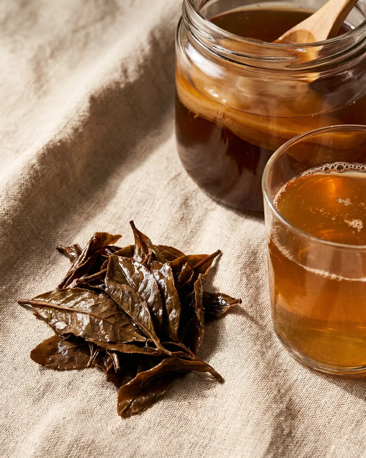 kombucha controindicazioni Foglie di tè umide accanto a un bicchiere di kombucha su una superficie di lino grezzo.