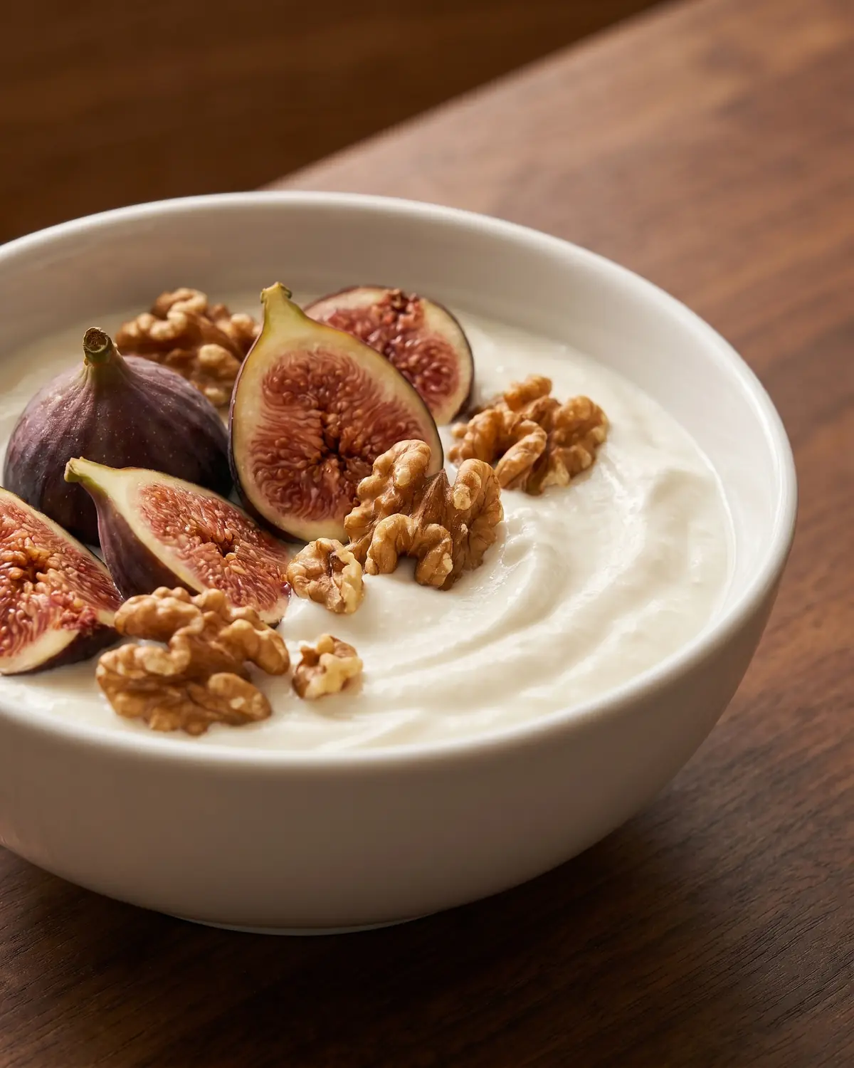 Yogurt in una ciotola bianca con fichi freschi e noci, visto di lato su legno scuro.