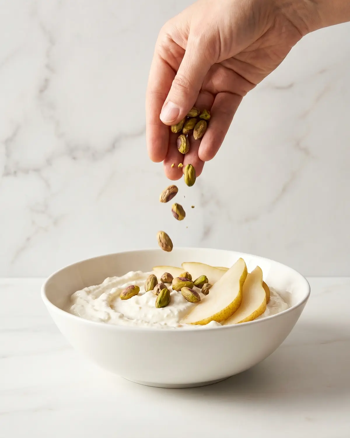 Mano che aggiunge pistacchi su yogurt con pera, in una ciotola su marmo bianco in verticale.